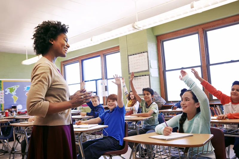 professora em sala de aula sorrindo para alunos com as maos levantadas